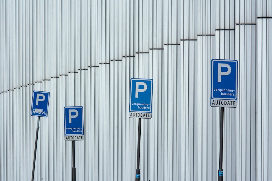 Four blue parking permit signs mounted on metal poles are aligned in front of a tall, light grey corrugated metal wall. The signs display a large white letter 'P' with additional text indicating parking permissions for loading and unloading, with the word 'AUTODATE' at the bottom. The signs are positioned at slightly different heights across the paved area, which may serve as a designated loading zone for house removals or furniture transport, as part of a home relocation process. The placement and design of the signs suggest they regulate parking for vehicles involved in moving services, aligning with the context of house removals and logistics support by companies such as Man with Van Highgate.