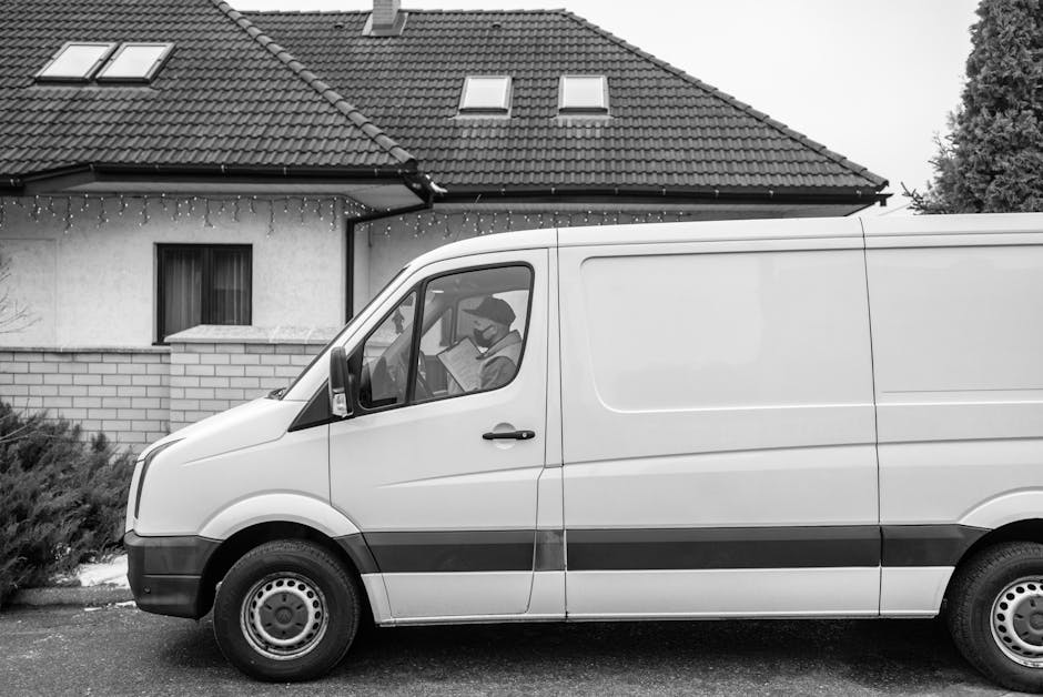 A white man with Van Highgate van parked on the street in front of a residential building with a tiled roof and small dormer windows. The van is positioned parallel to the curb, with the driver's side visible, showing a person wearing a face mask and reading a document inside through the window. The van's side door is closed, and it has a dark stripe running along the middle. Nearby, a driveway or loading area is visible, with shrubbery close to the building. The scene appears to capture part of the home relocation or furniture transport process, typical of professional removals and packing services, with the van ready for loading or unloading used items during a house move.