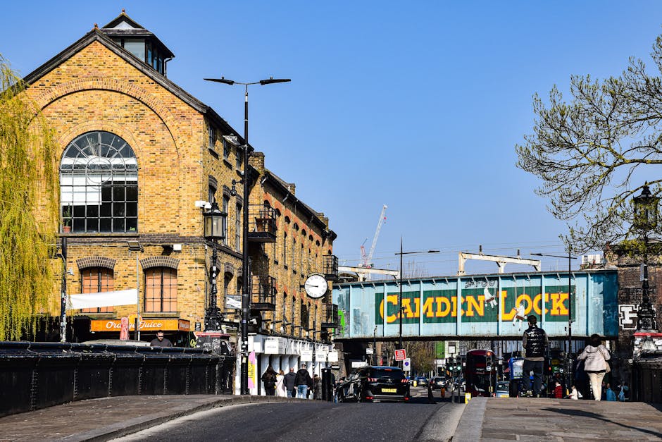 A busy street scene in Highgate, N6, featuring a large brick building with arched windows and a clock mounted on a black pole in front of it. To the right, a railway bridge with a blue painted sign reading 'CADDIN LOCK' spans across the street, indicating proximity to a local train station. Pedestrians are seen walking along the pavement, some carrying plastic bags or waiting, while cars and buses are parked or moving along the road. A few people are near the entrance of shops beneath the building, with some partially obscured by street furniture and streetlights. The area includes a variety of materials such as brick, metal, and glass, with trees lining parts of the street, adding greenery. The scene is well-lit with clear daytime sky overhead, reflecting typical urban activity involved in house removals and home relocation processes, especially relevant for end-of-lease clearance services addressed in the page about Camden Council waste rules.