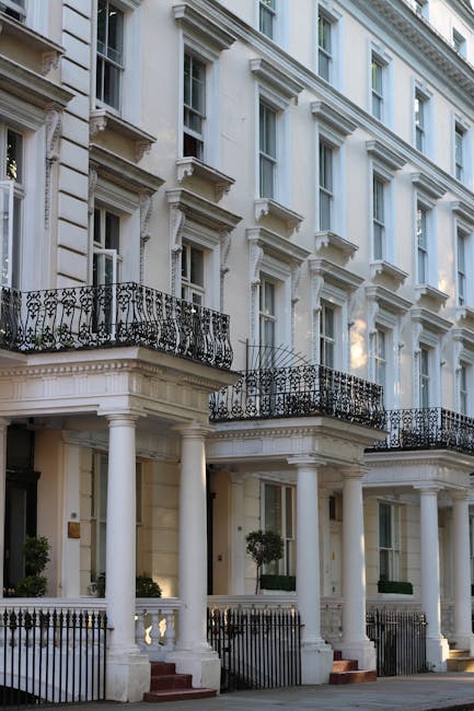 The image shows the exterior façade of a historic Georgian-style building in Highgate, characterized by tall white windows with decorative molding and black wrought-iron balconies. The ground floor features a porch supported by white columns, leading to the entrance door, with a small front garden enclosed by a black metal fence. Adjacent to the building, a moving van is parked on the pavement, partially visible on the right side of the image, with some cardboard boxes, wrapped furniture pieces, and packing materials like plastic wrap and blankets arranged nearby. The loading area of the van is open, indicating active loading or unloading of household items during a local house relocation. The scene is lit by natural daylight, and the surrounding architecture suggests a well-maintained residential area. This setup reflects the process of furniture transport and home relocation, in coordination with professional removals services such as those offered by Man with Van Highgate, as part of a detailed house move case study.