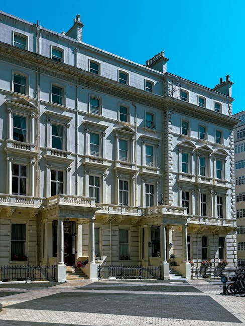 The exterior of a multi-storey white building with classical architectural details, including decorative columns, balustrades, and window pediments, under a clear blue sky. The ground floor features large windows and steps leading up to the entrances, with small potted plants near the doorways. The pavement in front is paved with light and dark tiles, and a black metal fence runs along the front. In the context of a home relocation, this building represents a residential property targeted for furniture transport and moving services, with the house likely being part of a house removal process carried out by Man with Van Highgate, who would manage the packing, loading, and transportation logistics involved in the move.