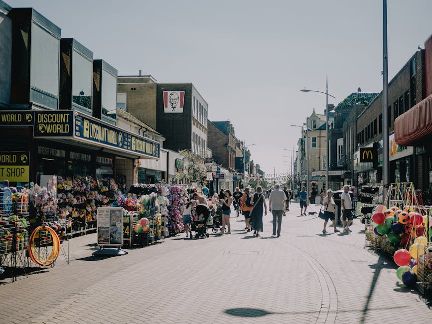 A busy outdoor shopping street on a clear day, with pedestrians walking along the pavement, some carrying shopping bags or pushing strollers. Small shops line both sides of the street, including a discount store with signage displaying 'DISCOUNT WORLD' and a fast food restaurant with a McDonald's logo. In front of the stores, there are displays of colorful items such as balloons, flowers, and outdoor merchandise arranged on metal racks or carts. The surrounding buildings are a mix of modern and older architectural styles, with large windows and signage. The road is free of vehicles, and the scene is illuminated by street lamps and natural sunlight, suggesting a daytime setting. This environment depicts a typical urban commercial area, relevant to services like house and shop removals or relocation, as provided by Man with Van Highgate.
