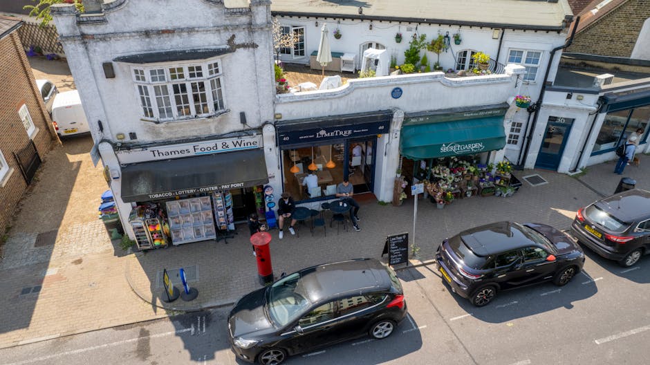 An aerial view of a street scene showing a white commercial building with multiple storefronts, including Thames Food & Wine with a striped awning, a restaurant called La Petite and a flower shop with a green canopy. In front of these shops, three cars are parked along the curb, with the nearest being a black hatchback. The pavement area in front of the shops has a few individuals sitting on benches, with some engaging in conversation, and others standing. A red post box is situated on the sidewalk near the benches. The street scene captures the typical elements of a small retail environment, featuring display windows, awnings, and outdoor seating, with surrounding residential and commercial buildings. This image relates to the context of house removals and moving logistics, highlighting the need for careful planning and transportation of furniture and packing materials in and around small shop office relocations, as provided by Man with Van Highgate.
