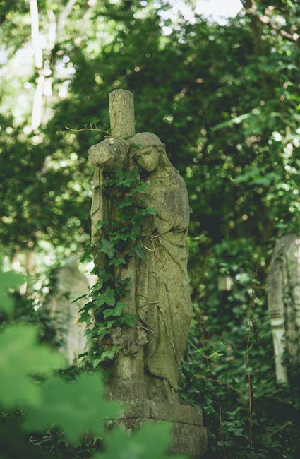 A weathered stone statue of a religious figure standing outdoors among greenery, with a tall stone cross behind it. The statue depicts a person dressed in robes, holding an object close to their chest