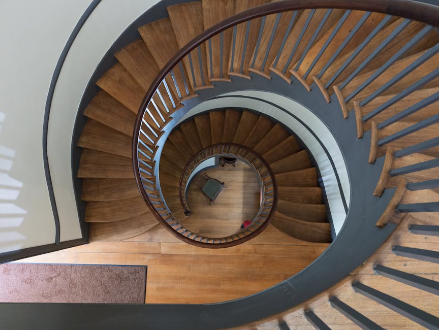 An overhead view of a wooden spiral staircase inside a residential property, captured from the top looking down towards the ground floor. The staircase features curved wooden steps with a dark grey painted riser, and a smooth, rounded wooden handrail following the spiral design. The balustrade consists of evenly spaced vertical wooden balusters. The cladding around the staircase is painted in a neutral light colour, contrasting with the warm tone of the finished wooden steps. At the bottom of the staircase, a small green upholstered chair with wooden legs is placed on the wooden floor, adjacent to a grey floor mat near the entrance or hallway area. The interior space is well-lit, with natural light possibly coming from an unseen window or adjacent room, creating shadows on the steps and balustrade. This image supports home relocation services by illustrating the challenges of navigating furniture through tight staircases, which [COMPANY_NAME], such as Man with Van Highgate, may encounter during removal or furniture transport within a property or between locations.