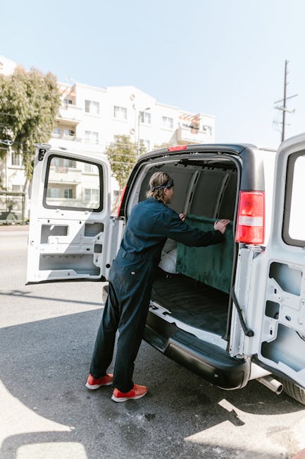 A person wearing a dark blue work jumpsuit with red shoes is loading a large flat-screen television wrapped in green anti-scratch padding into the cargo area of a white van parked on a residential str