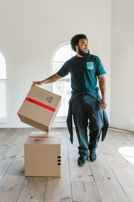A person wearing a dark blue work jumpsuit with red shoes is loading a large flat-screen television wrapped in green anti-scratch padding into the cargo area of a white van parked on a residential str
