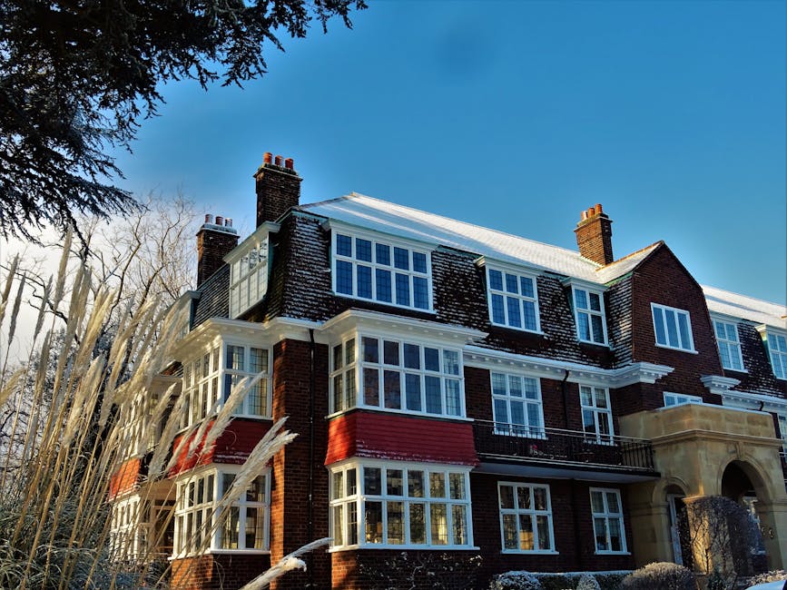 Photograph of a large multi-story residential building with a brick facade, situated on Swains Lane in Highgate. The building features numerous white-framed sash windows, some with small balconies, and has a classical architectural style with decorative stonework around the entrance. The roof is partially covered with snow, and several tall brick chimneys are visible at the top. Surrounding the property, there are leafless trees and tall ornamental grasses, indicating a winter season. The sky above is clear and blue. This image, captured during daylight, exemplifies a typical period property suitable for house removals and furniture transport, which [COMPANY_NAME] may assist with during home relocation or packing and moving services.