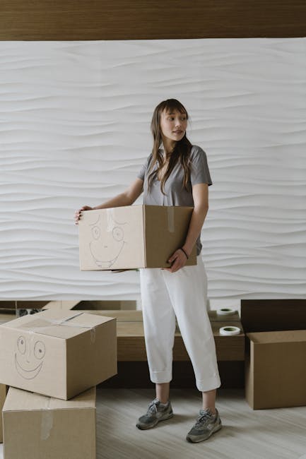 A woman holding a cardboard box with a hand-drawn smiley face, standing in front of a textured white wall inside a room. She is dressed in casual grey T-shirt, light-colored trousers, and grey sneakers. In front of her are two additional cardboard boxes, one on the floor and another resting on her arm, also featuring smiley face drawings. The setting appears to be part of a home relocation or packing process, with the boxes likely prepared for moving. The environment includes a wooden floor and a small dark wooden bench with a few stationary items on it, indicating an organized space. The image, associated with house moves and removal services, reflects the packing and handling phase of furniture transport and home relocation, with [COMPANY_NAME] providing professional moving support, as indicated by the website details.